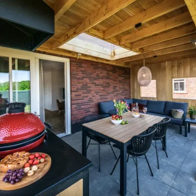 Ein modernes Ferienhaus mit großer Terrasse, Esstisch und einem auffälligen roten Grill lädt zum gemütlichen Beisammensein im Freien ein.
