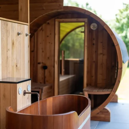 Außenansicht eines rustikalen Wellnessbereichs mit hölzerner Ausstattung, einer freistehenden Holzbadewanne und einer runden Fasssauna mit Glastür und Blick auf die grünen Außenflächen.